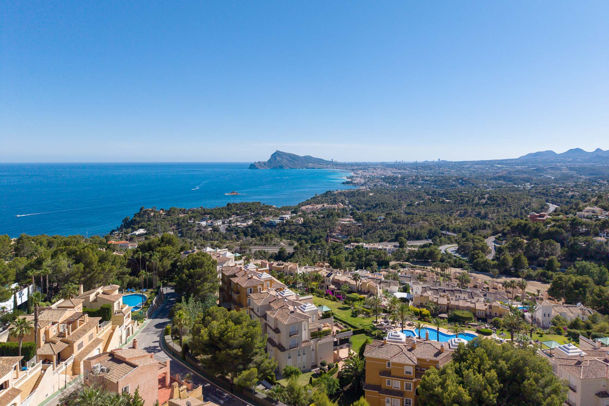 Vue aérienne de la côte d'Altea Hills, avec la mer Méditerranée, le Peñón de Ifach et la silhouette de Benidorm au loin. Toits en terre cuite et piscines se distinguent parmi la végétation luxuriante.