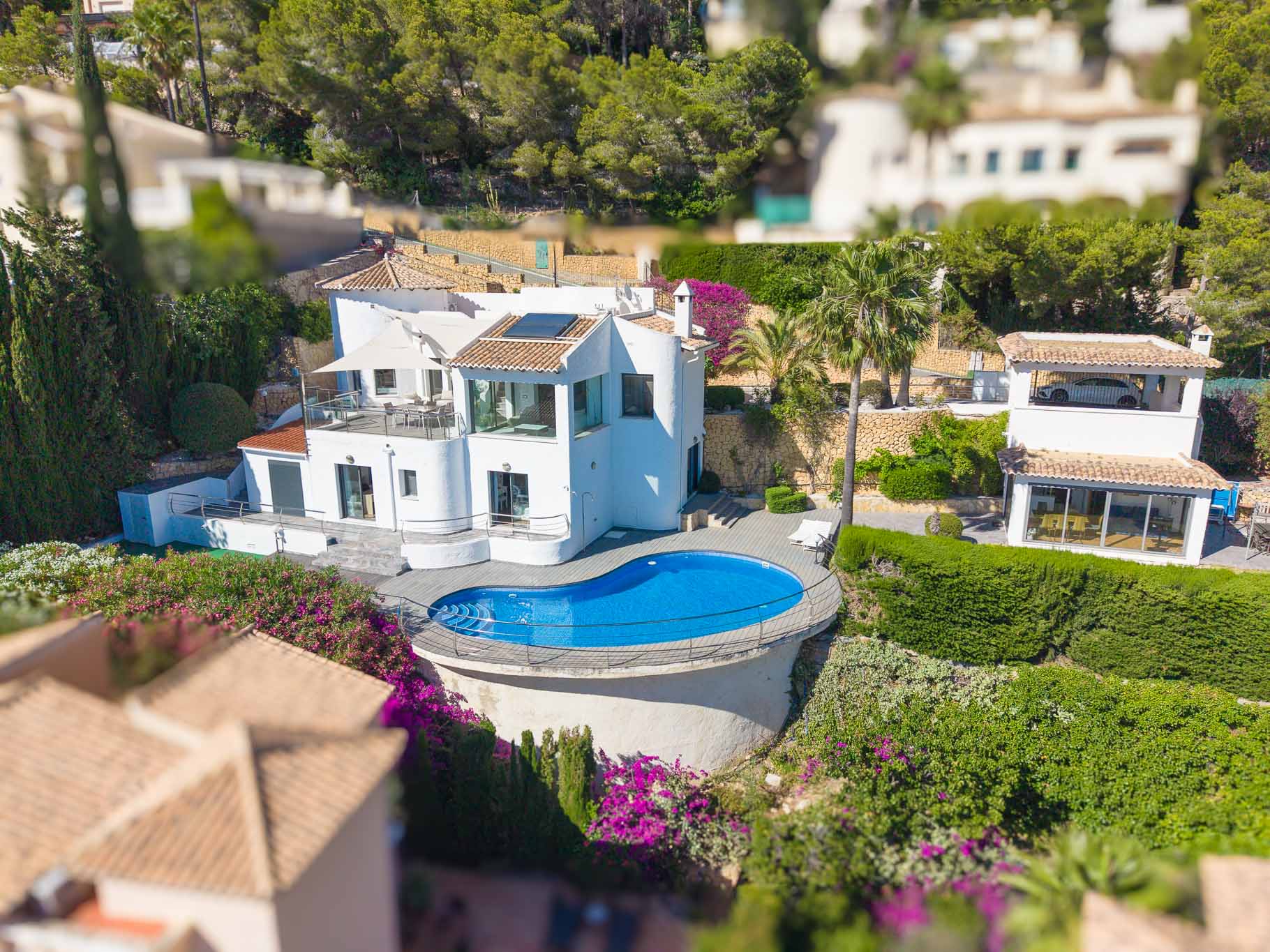 Vue aérienne de la villa blanche, présentant une piscine de forme libre avec terrasse en bois et garde-corps. On y aperçoit plusieurs terrasses et des jardins méditerranéens luxuriants.