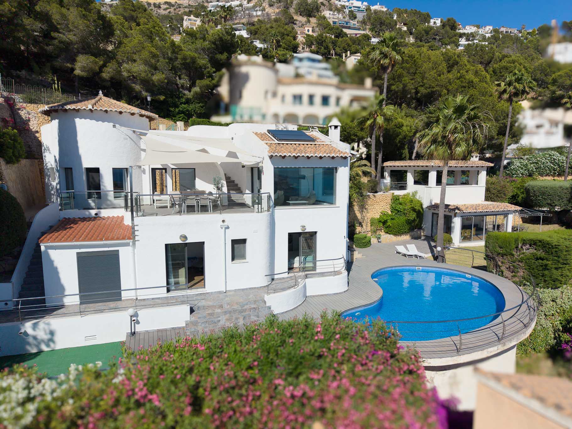 Vue aérienne de la villa principale, avec une vaste terrasse supérieure ombragée, une piscine en forme de rein avec sa plage et une dépendance séparée, le tout niché dans un paysage méditerranéen verdoyant.