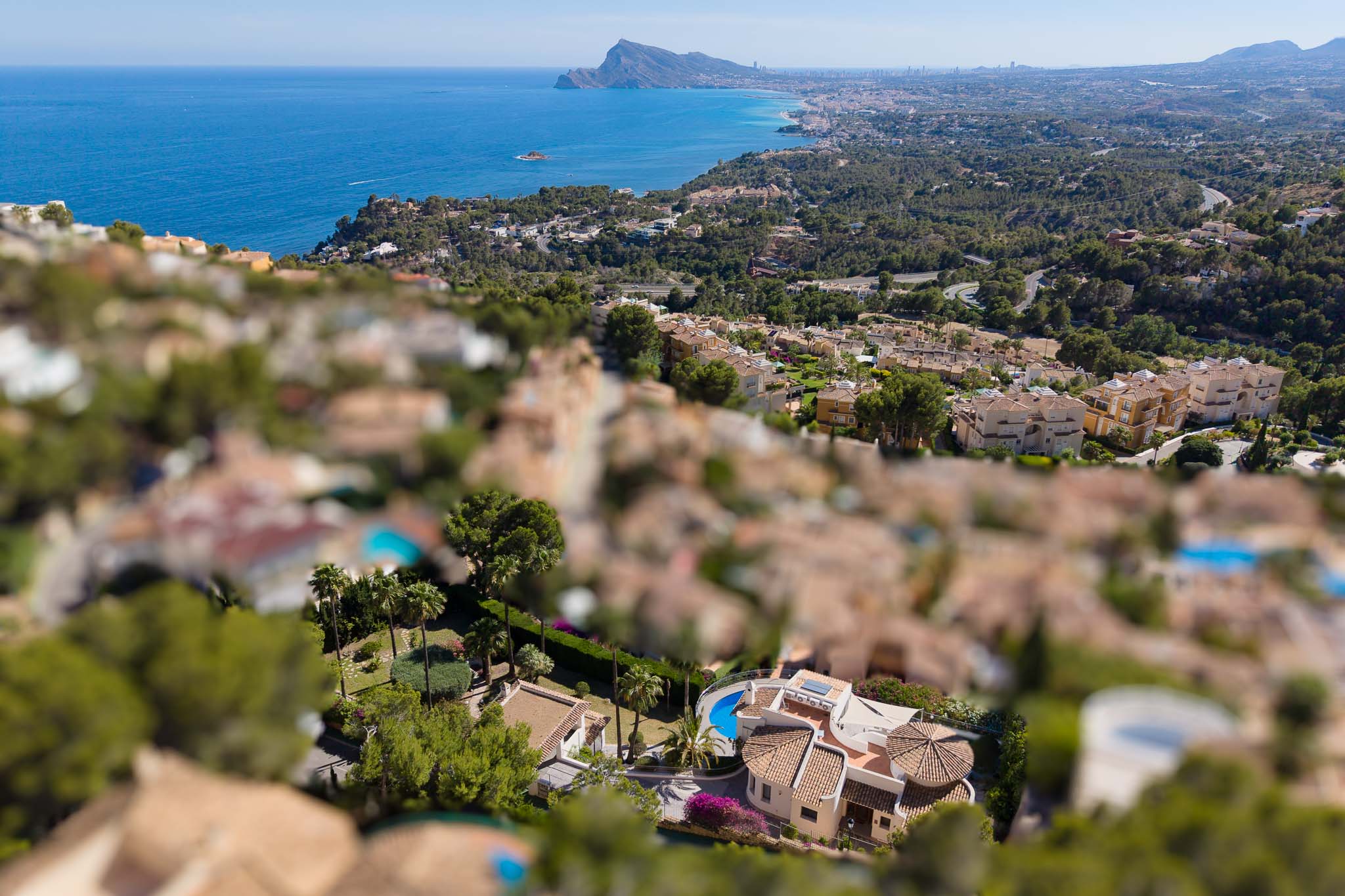 Vue aérienne d'une villa à Altea Hills avec piscine incurvée, toits en terre cuite et jardins luxuriants, sur fond de mer Méditerranée et de côte.