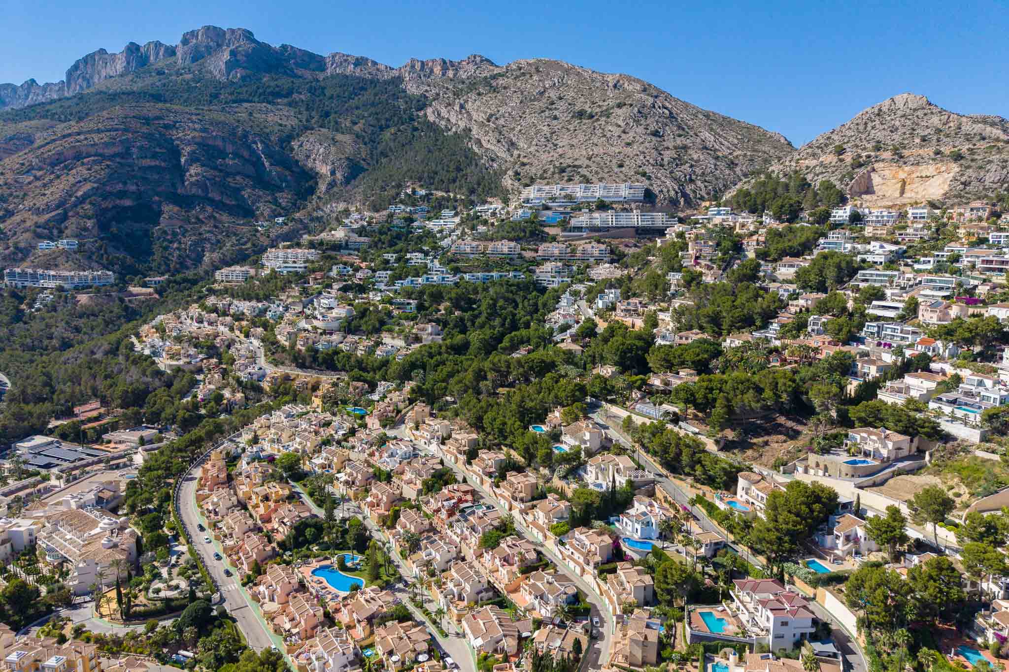 Vue aérienne panoramique d'Altea Hills, présentant de nombreuses villas avec piscines sur la colline verdoyante, adossées à des montagnes imposantes.