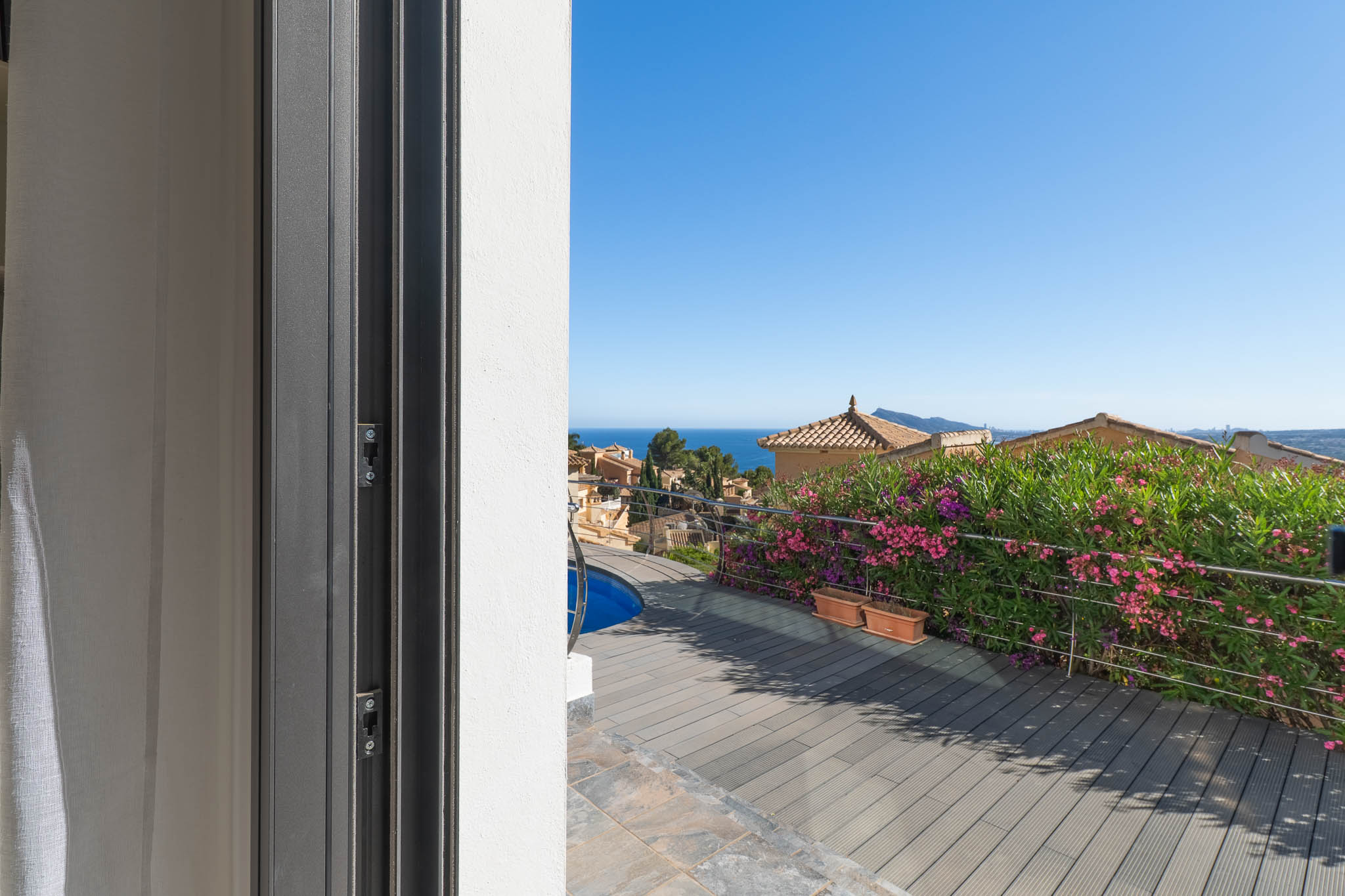 Vue depuis la porte coulissante sur la terrasse en composite, aperçu de la piscine et garde-corps en acier. La mer Méditerranée et les toits traditionnels sont visibles en arrière-plan.