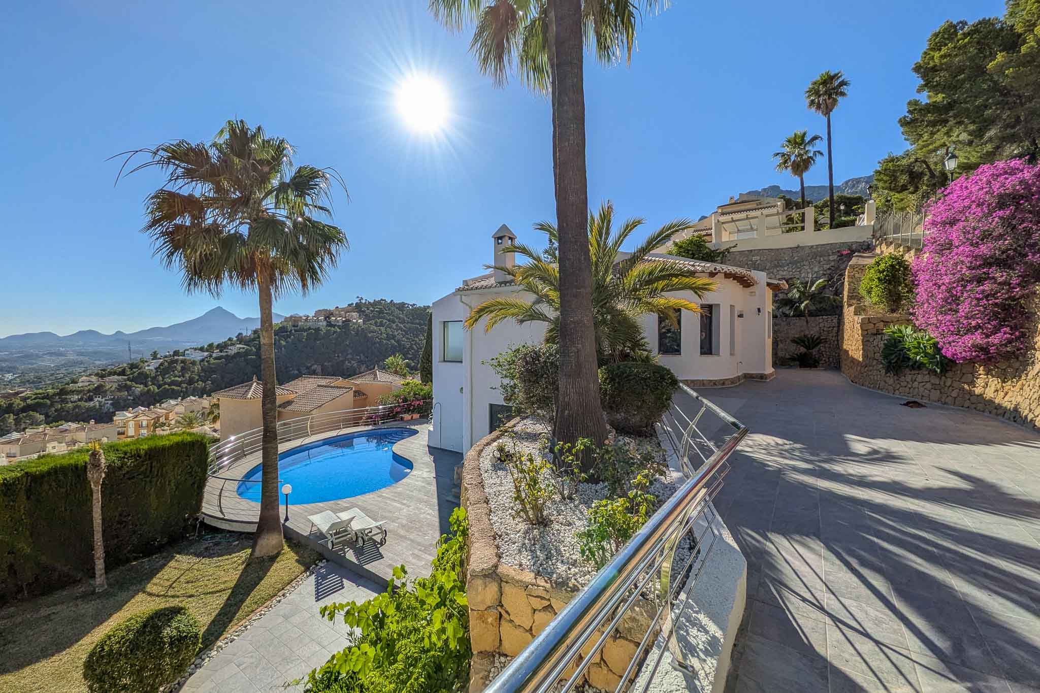 Vue extérieure de la villa avec piscine en forme de rein, entourée de palmiers et d'un jardin soigné. L'allée pavée monte le long de murs en pierre et de bougainvilliers, offrant des vues sur les montagnes.