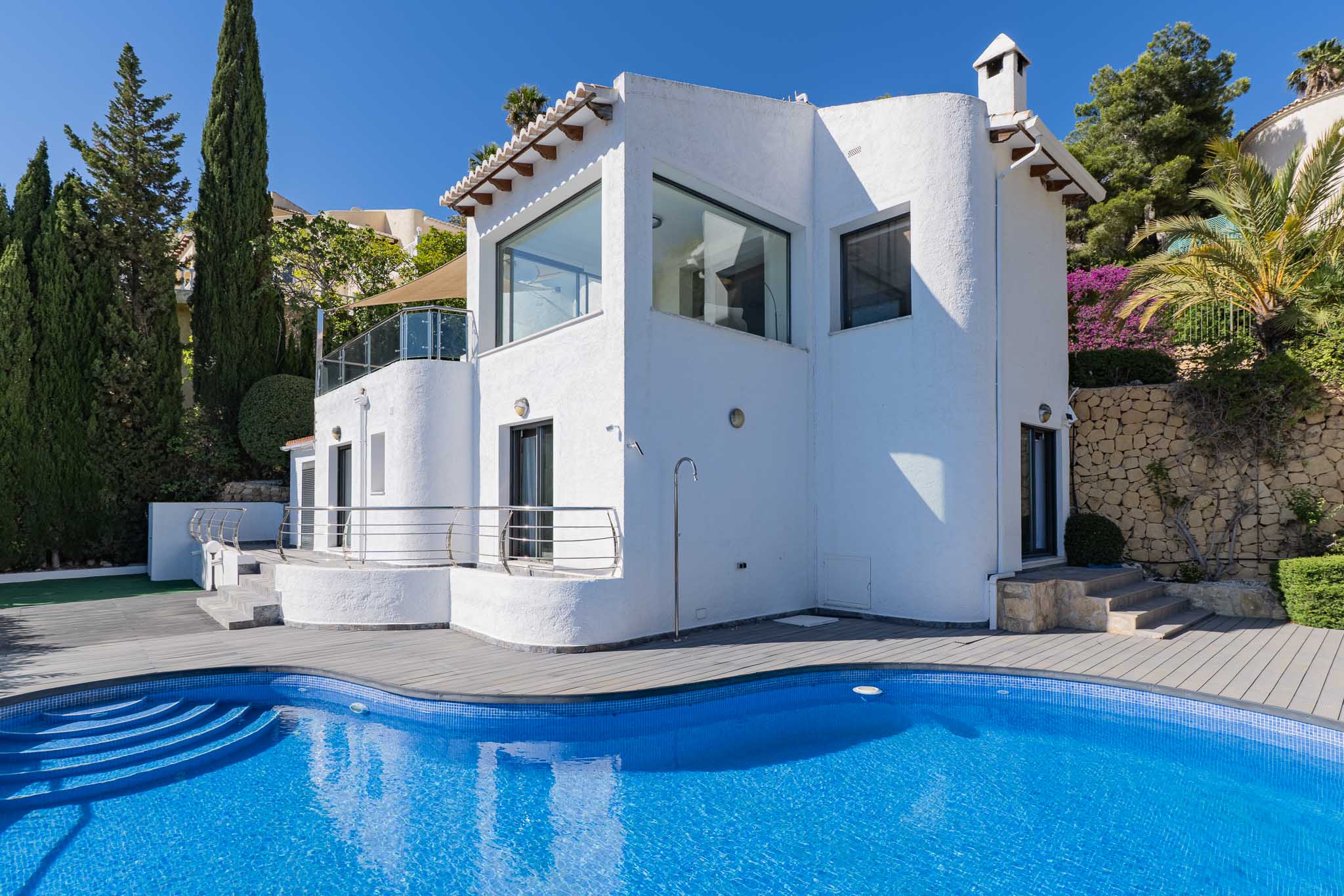 Vue extérieure de la villa, présentant une piscine courbe en mosaïque bleue avec terrasse composite grise, douche extérieure et flore méditerranéenne.