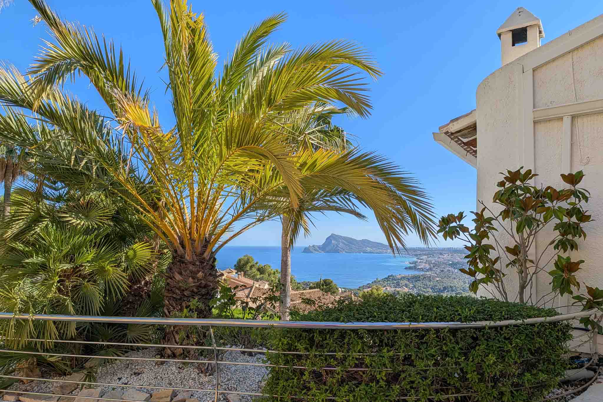 Vue mer panoramique depuis une terrasse, encadrée par des palmiers luxuriants. Balustrade en acier inoxydable, haie verte et littoral avec le Peñón de Ifach.