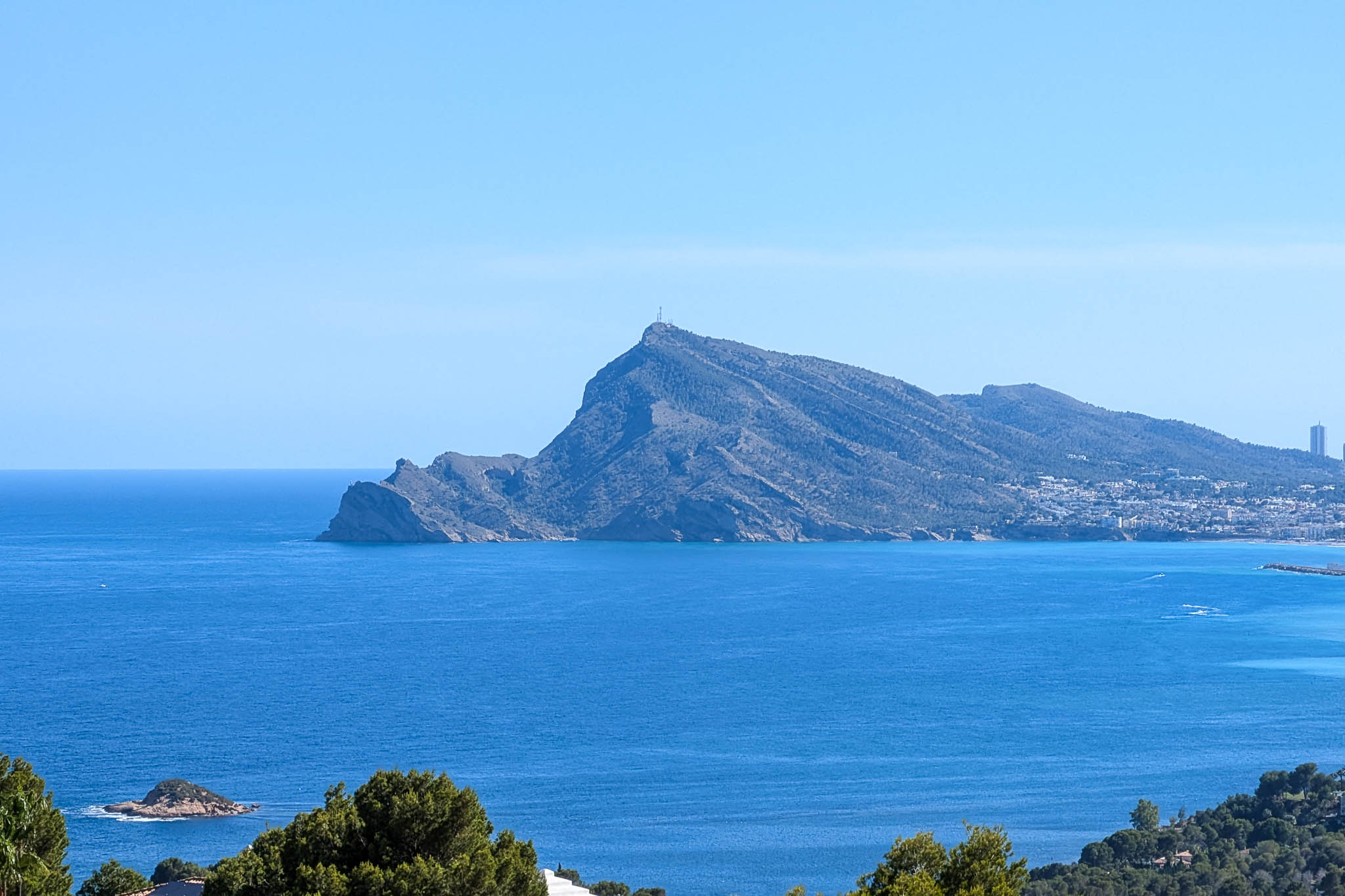 Vue panoramique sur la Méditerranée, avec le profil imposant de la Sierra Helada, une ville côtière lointaine et de petits îlots rocheux sous un ciel azur.
