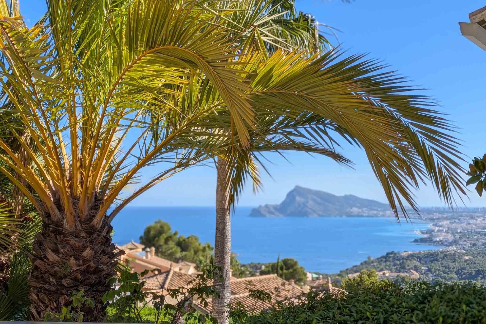 Vue panoramique sur la mer Méditerranée, encadrée par des palmiers, avec les toits d'Altea Hills et le Peñón de Ifach au loin sous un ciel bleu.