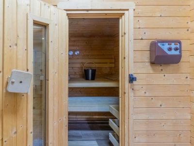 Accès au sauna en bois clair, avec ses bancs intérieurs, un seau et une louche, ainsi qu'un panneau de contrôle mural externe.