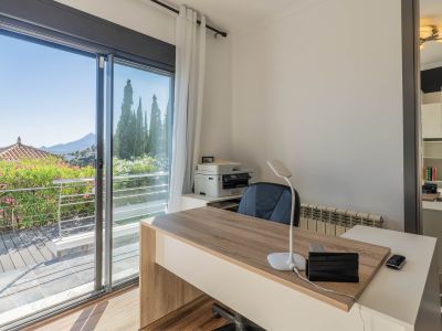 Bureau lumineux avec un bureau moderne en bois, une grande porte coulissante vitrée s'ouvrant sur une terrasse verdoyante et des vues panoramiques sur la montagne. Un miroir reflète une étagère fonctionnelle.