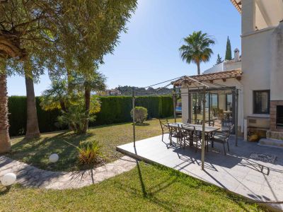 Cet espace repas extérieur comprend un patio carrelé sous une pergola métallique, parfait pour les repas en plein air. Il est complété par un barbecue intégré et un jardin luxuriant avec palmiers et haie.