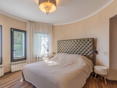 Chambre avec grand lit et tête de lit capitonnée. Mur incurvé avec fenêtres à cadre sombre et voilages clairs. Parquet, radiateurs et luminaire de plafond moderne.