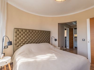 Chambre principale avec tête de lit capitonnée, mur incurvé et lumière naturelle filtrée par des voilages, complétée par des portes en bois.
