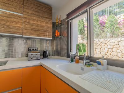 Cuisine moderne avec comptoir en quartz blanc, armoires basses orange vif et unités supérieures en bois veiné. Fenêtre offrant une vue sur un mur de pierre et la végétation.