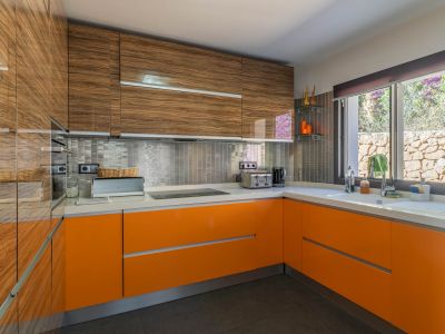 Cuisine moderne avec meubles hauts en bois veiné brillant et bas laqués orange vif. Le plan de travail blanc, la crédence en mosaïque métallique et la fenêtre donnant sur un mur de pierre sont visibles.