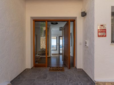 Entrée principale avec porte pivotante en bois et panneau vitré, ouvrant sur un intérieur lumineux aux parquets et vues mer. Murs extérieurs texturés et carrelage foncé.
