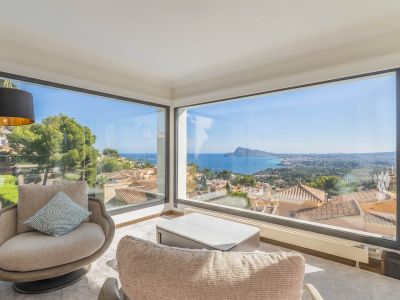 Espace salon avec de larges baies vitrées d'angle offrant une vue panoramique sur la mer et la côte. Deux fauteuils et une table basse moderne créent un lieu lumineux.