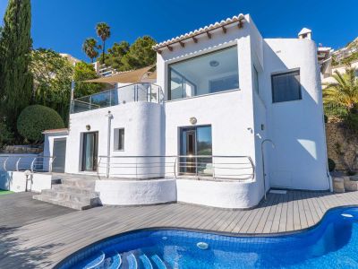 Extérieur de la villa blanche avec grandes baies vitrées et terrasses à balustrades en verre. La piscine en mosaïque bleue et le pont en composite gris sont entourés de cyprès et de palmiers.