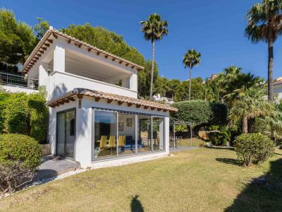 Façade blanche de la villa avec tuiles traditionnelles et grandes baies vitrées. Le jardin plat comprend une pelouse verdoyante et un patio avec pergola, entouré de palmiers et arbustes.