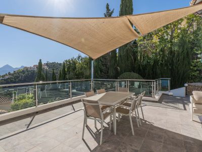 Grande terrasse extérieure ombragée par une voile solaire, meublée d'une table à manger et bordée d'une balustrade en verre, avec vue panoramique sur les montagnes.