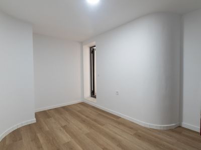 Pièce lumineuse avec sol en lames claires, murs blancs et une paroi courbe distinctive. Présente une ouverture verticale à cadre sombre et seuil en pierre.