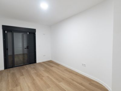 Pièce lumineuse avec un sol stratifié imitation bois clair et des murs blancs. Une grande porte coulissante en verre gris anthracite avec stores intégrés mène à l'extérieur.