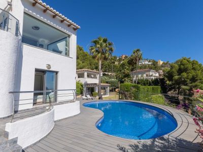 Piscine courbe en mosaïque bleue avec terrasse en composite gris, jouxtant une façade blanche dotée d'une fenêtre panoramique et d'un balcon à balustrade en acier.