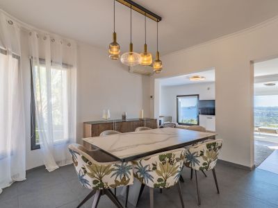 Salle à manger moderne avec table effet marbre et chaises à motif tropical. Un lustre linéaire éclaire l'espace, qui s'ouvre sur des vues panoramiques sur la mer depuis les pièces adjacentes.