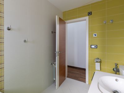 Salle de bain moderne avec carrelage mural vert anis et beige texturé, agrémenté d'une bande mosaïque. Elle est équipée d'un lavabo contemporain et d'une porte en bois.