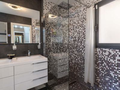 Salle de bain moderne avec double vasque, meuble blanc et miroir éclairé. La douche vitrée est revêtue de mosaïque grise et inclut une fenêtre intégrée.