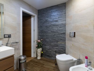 Salle de bain moderne avec mur d'accent en pierre empilée foncée, carrelage beige clair et sol imitation bois. Comprend toilettes, bidet et meuble-lavabo.