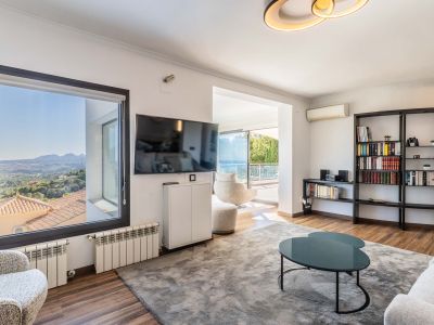 Salon lumineux offrant des vues panoramiques sur la montagne depuis la fenêtre et un accès à la terrasse avec vue sur mer. Il présente un sol en bois, un grand tapis gris et une bibliothèque moderne.
