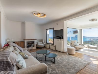 Salon moderne avec canapé clair, cheminée et larges baies vitrées offrant des vues panoramiques sur la mer et la ville. Le mobilier contemporain illumine l'espace.
