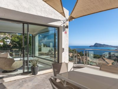 Terrasse extérieure aménagée avec table et chaises, sous une voile d'ombrage. Elle offre des vues panoramiques sur la mer et la côte via une balustrade en verre, ouvrant sur un intérieur lumineux.