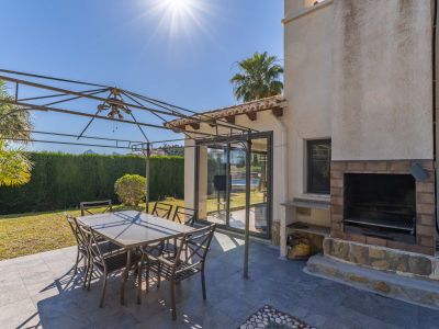 Terrasse extérieure carrelée en gris foncé, avec pergola métallique et table à manger. Un barbecue intégré en brique et pierre et des portes vitrées coulissantes sont visibles.