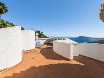 Terrasse sur le toit avec des carreaux de terre cuite et des murs blancs texturés, offrant une vue panoramique sur la mer et la côte. Une chaise longue bleue invite à la détente.