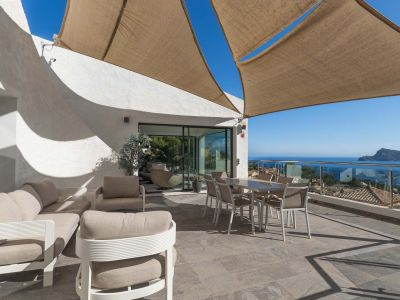 Vaste terrasse extérieure avec salon et salle à manger sous voile d'ombrage, offrant une vue panoramique sur la mer et la côte via une balustrade en verre.