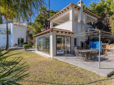 Vaste terrasse pavée avec salle à manger extérieure sous pergola métallique, jouxtant une pièce vitrée et un jardin méditerranéen entretenu.