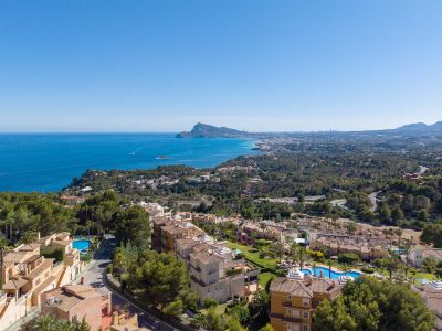 Vue aérienne de la côte d'Altea Hills, avec la mer Méditerranée, le Peñón de Ifach et la silhouette de Benidorm au loin. Toits en terre cuite et piscines se distinguent parmi la végétation luxuriante.