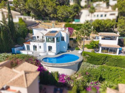 Vue aérienne de la villa blanche, présentant une piscine de forme libre avec terrasse en bois et garde-corps. On y aperçoit plusieurs terrasses et des jardins méditerranéens luxuriants.