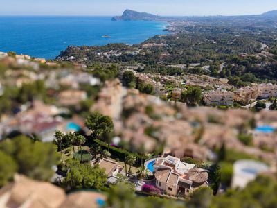 Vue aérienne d'une villa à Altea Hills avec piscine incurvée, toits en terre cuite et jardins luxuriants, sur fond de mer Méditerranée et de côte.