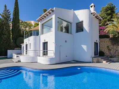 Vue extérieure de la villa, présentant une piscine courbe en mosaïque bleue avec terrasse composite grise, douche extérieure et flore méditerranéenne.
