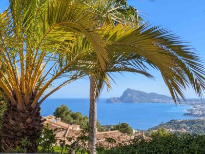 Vue panoramique sur la mer Méditerranée, encadrée par des palmiers, avec les toits d'Altea Hills et le Peñón de Ifach au loin sous un ciel bleu.