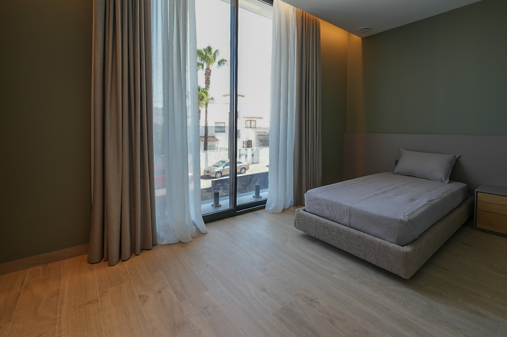 Chambre avec lit simple, parquet clair et murs vert olive. Les grandes baies vitrées avec doubles rideaux offrent une vue sur la rue et les palmiers. Chambre avec lit simple, parquet clair et murs vert olive. Les grandes baies vitrées avec doubles rideaux offrent une vue sur la rue et les palmiers.