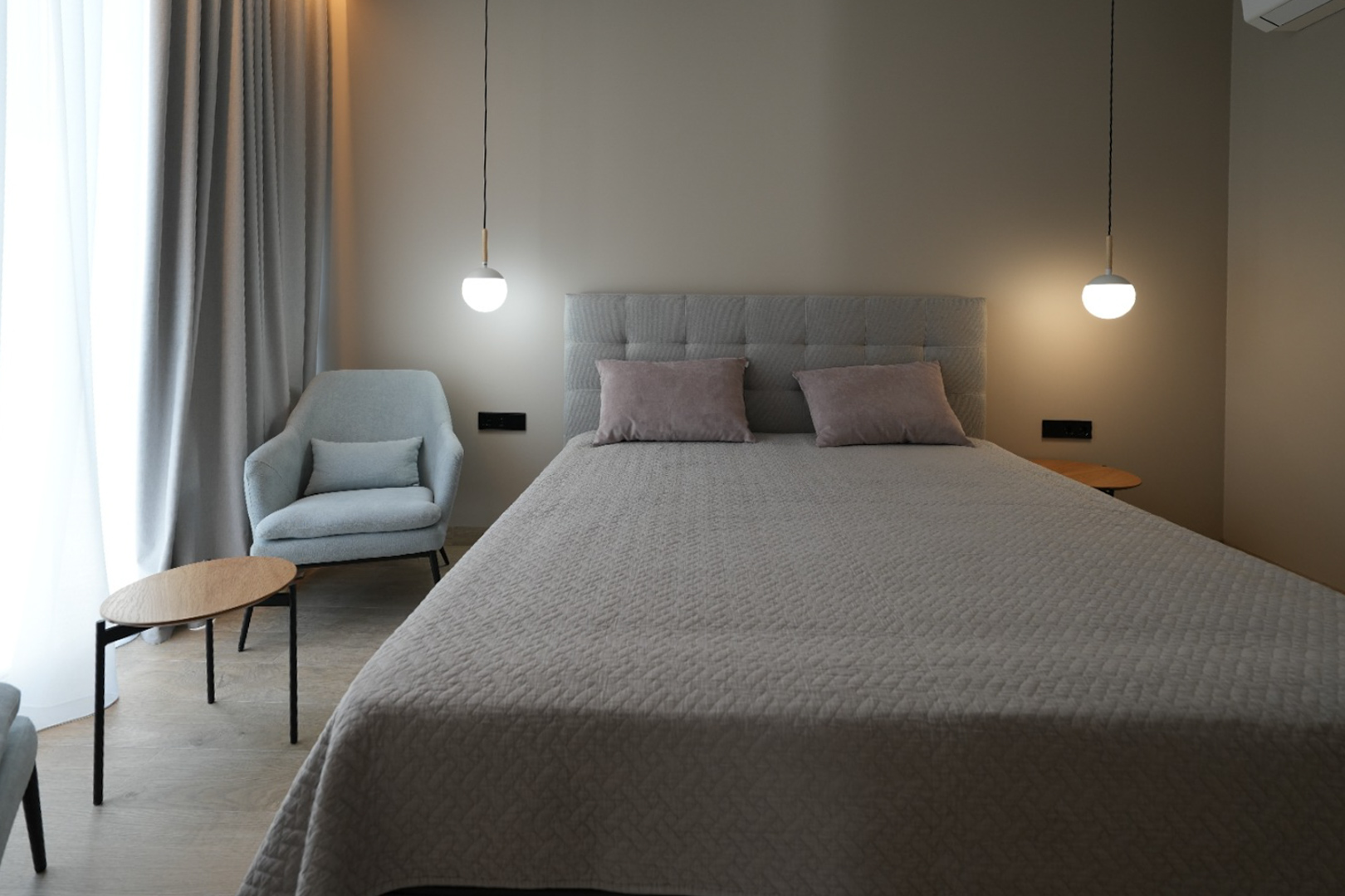 Chambre élégante avec un lit capitonné gris, deux suspensions modernes et un fauteuil confortable. Les rideaux clairs et le sol effet bois créent une ambiance sereine. Chambre élégante avec un lit capitonné gris, deux suspensions modernes et un fauteuil confortable. Les rideaux clairs et le sol effet bois créent une ambiance sereine.