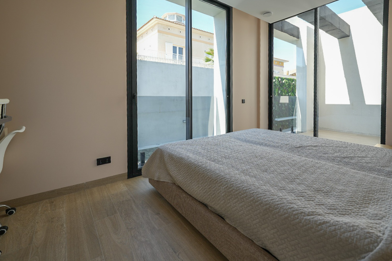 Chambre lumineuse avec parquet clair et grandes baies vitrées coulissantes à cadre noir, offrant une vue sur une maison voisine et un mur végétal extérieur. Chambre lumineuse avec parquet clair et grandes baies vitrées coulissantes à cadre noir, offrant une vue sur une maison voisine et un mur végétal extérieur.