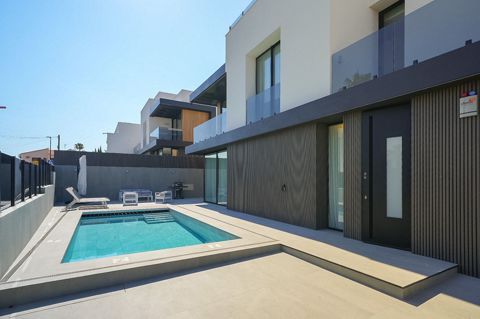 Extérieur de villa moderne avec piscine rectangulaire et escaliers intégrés, entourée d'un dallage clair. Façade du rez-de-chaussée avec revêtement à lattes sombres et porte d'entrée distinctive. Extérieur de villa moderne avec piscine rectangulaire et escaliers intégrés, entourée d'un dallage clair. Façade du rez-de-chaussée avec revêtement à lattes sombres et porte d'entrée distinctive.