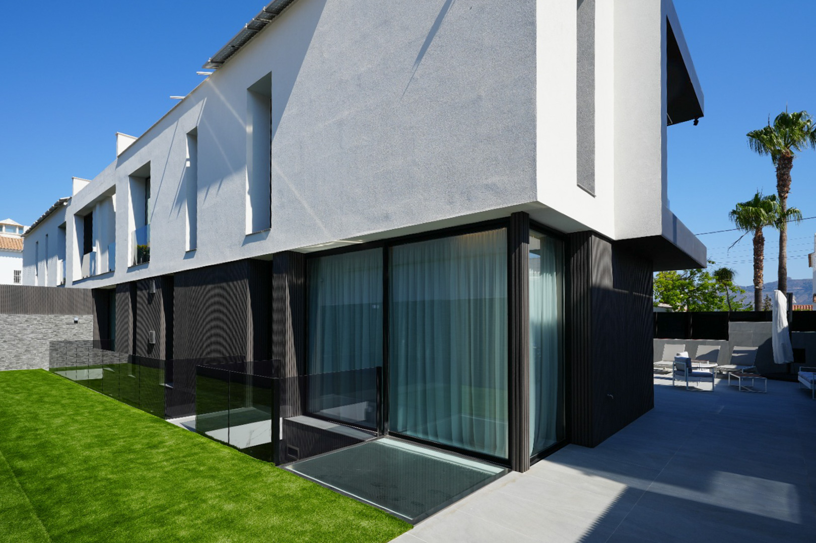 Façade moderne avec textures contrastées blanc/gris et sombre, grandes baies vitrées, pelouse artificielle et terrasse carrelée aménagée avec mobilier extérieur. Façade moderne avec textures contrastées blanc/gris et sombre, grandes baies vitrées, pelouse artificielle et terrasse carrelée aménagée avec mobilier extérieur.