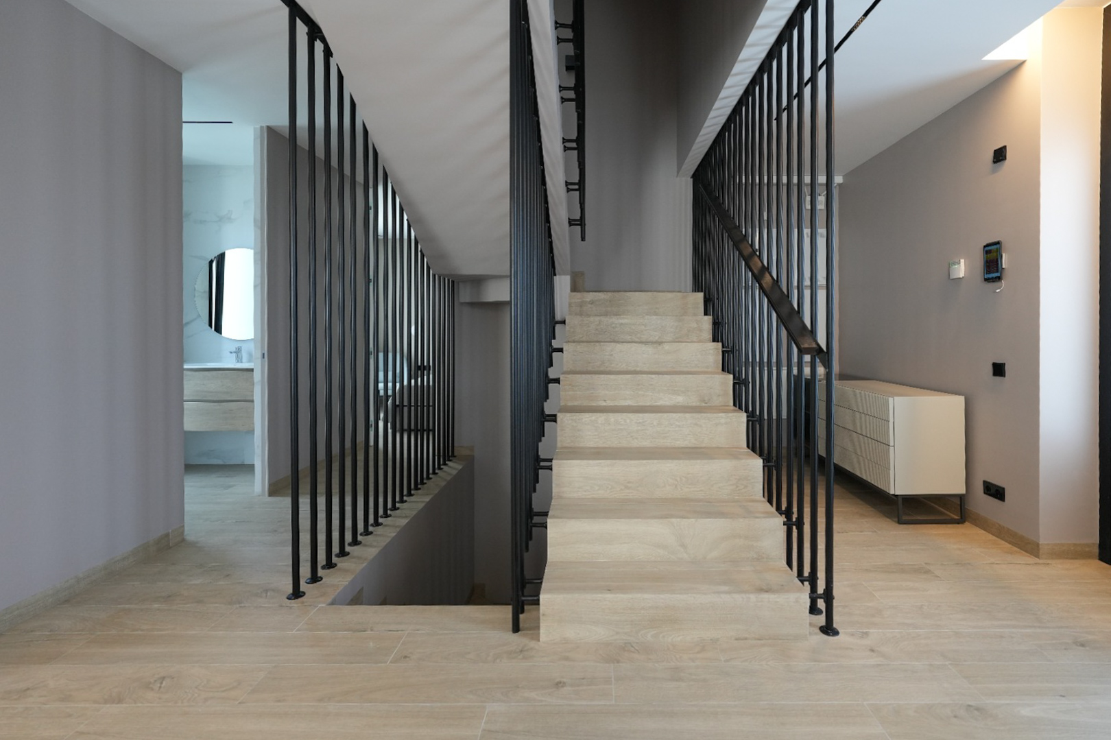 Intérieur moderne avec escalier en bois clair et balustrade verticale noire épurée. Un panneau de contrôle domotique est visible, menant à une salle de bain avec miroir rond. Intérieur moderne avec escalier en bois clair et balustrade verticale noire épurée. Un panneau de contrôle domotique est visible, menant à une salle de bain avec miroir rond.
