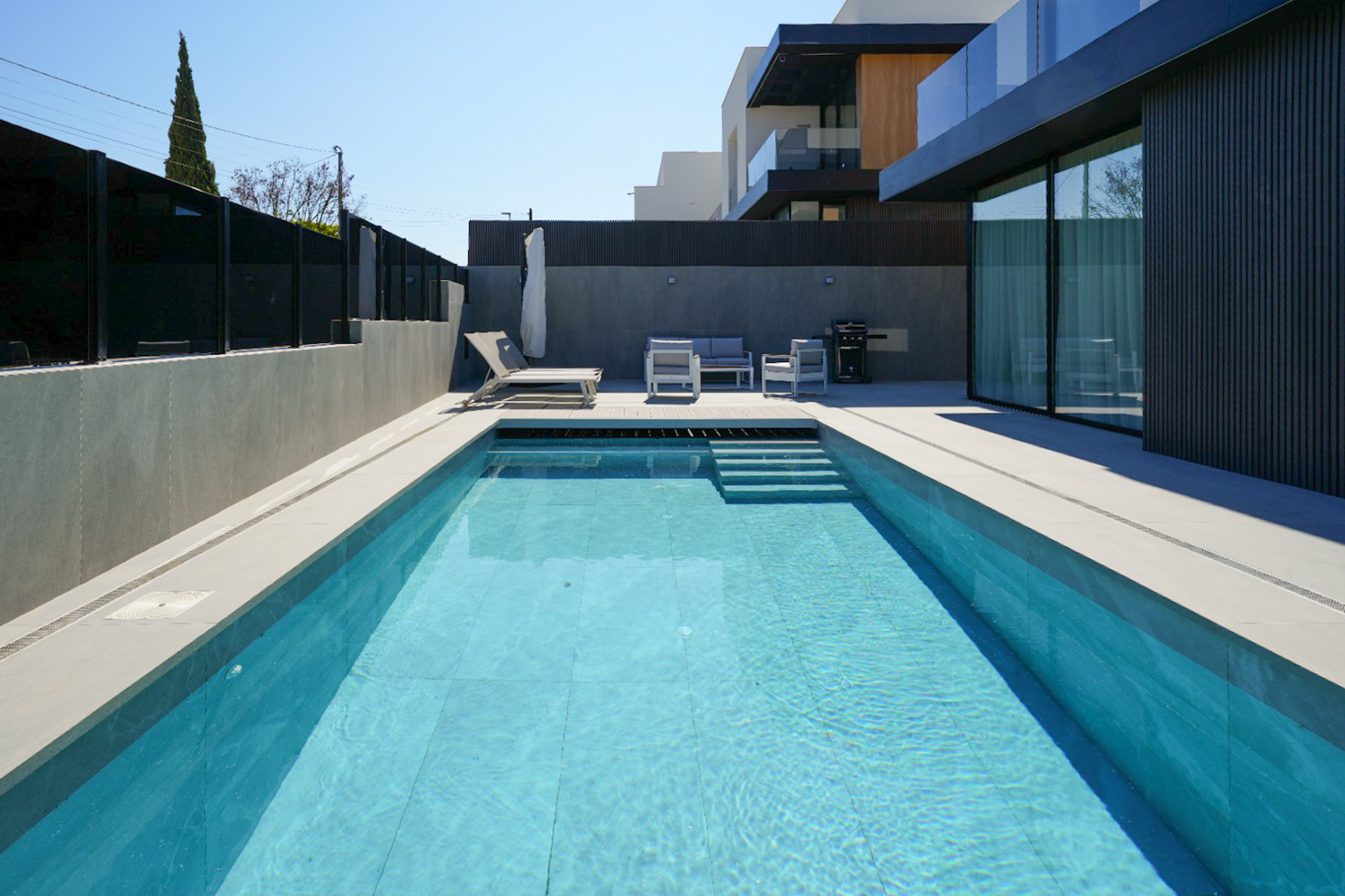 L'espace extérieur comprend une piscine rectangulaire aux carreaux bleus clairs et marches, bordée d'un dallage gris clair. Un salon de jardin moderne et une chaise longue invitent à la détente près de la façade épurée. L'espace extérieur comprend une piscine rectangulaire aux carreaux bleus clairs et marches, bordée d'un dallage gris clair. Un salon de jardin moderne et une chaise longue invitent à la détente près de la façade épurée.