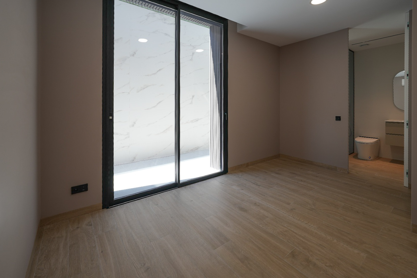 Pièce spacieuse avec sol carrelé imitation bois et murs clairs. De grandes portes coulissantes vitrées noires mènent à une terrasse extérieure carrelée de marbre blanc. Salle de bain intégrée visible. Pièce spacieuse avec sol carrelé imitation bois et murs clairs. De grandes portes coulissantes vitrées noires mènent à une terrasse extérieure carrelée de marbre blanc. Salle de bain intégrée visible.